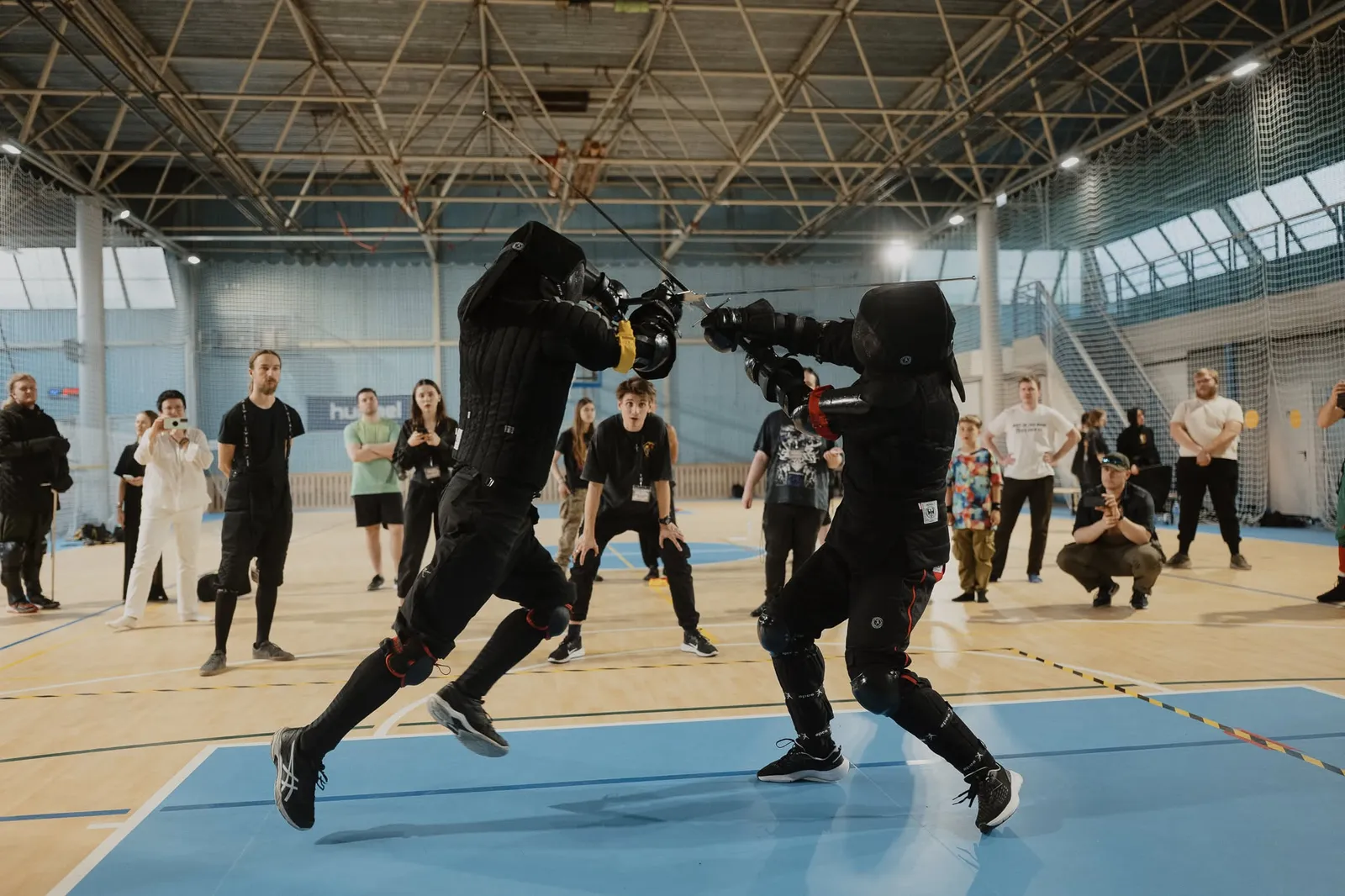 Ilgųjų kalavijų sparingas sporto salėje, aplink stebintys klubo nariai
