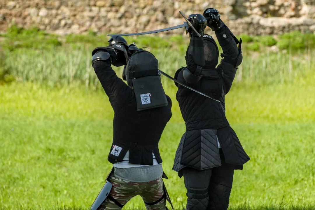Du fechtuotojai susikerta ilgaisiais kalavijais pievoje prie senos mūrinės sienos